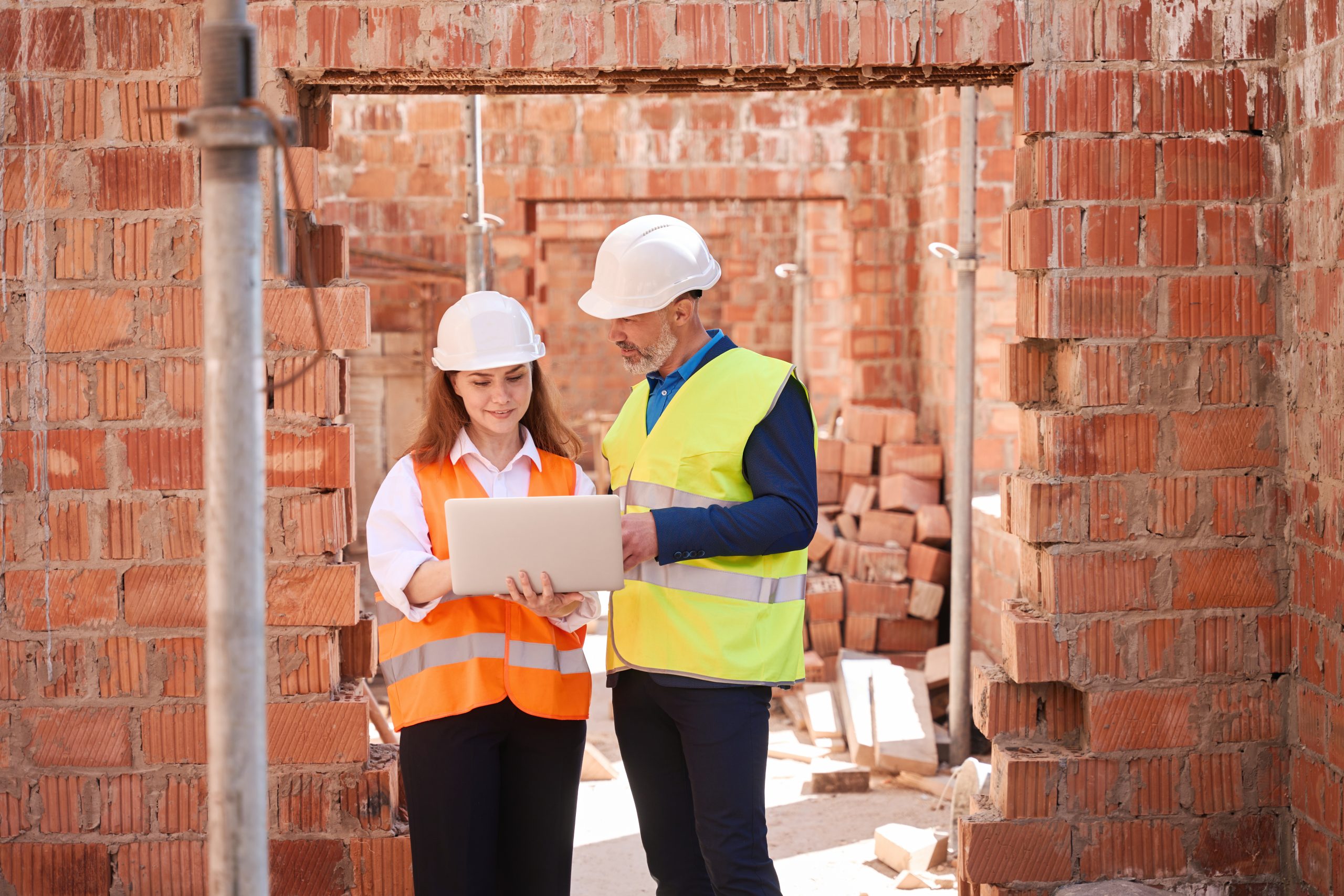 Building engineers in hardhats checking 3d building layout on laptop, standing inside building under construction, discussing design